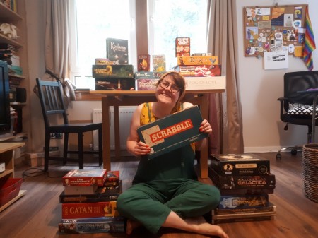Image of Jenny sitting cross-legged on the floor in front of a dining table piled with board games, surrounded by two piles of board games including Catan, Carcassone, Pandemic and others. She is holding an older version of the game Scrabble in her hands.